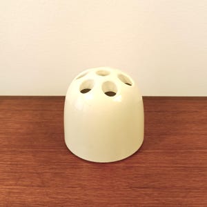 Peut inclure: Vase à bourgeon en céramique crème avec six ouvertures circulaires sur le dessus. Le vase a une forme arrondie en forme de dôme et repose sur une surface en bois. Le vase est de couleur neutre et a une finition brillante.