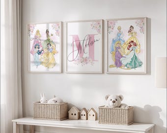 May include: Three framed art prints featuring watercolor illustrations of Disney princesses. The center print displays the name "Madison" over a pink letter "M". The other two prints showcase princesses in colorful gowns, surrounded by floral accents. The prints are displayed above a white table.
