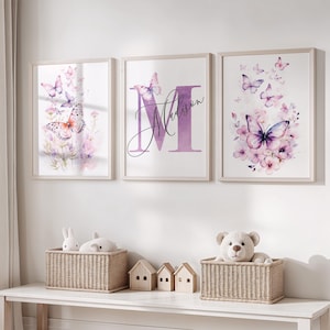May include: Three framed butterfly art prints in pastel shades. The prints depict watercolour butterflies and floral designs. The central print features the letter "M" and the name "Madison". The prints are displayed above a white table with wicker baskets and miniature wooden houses.