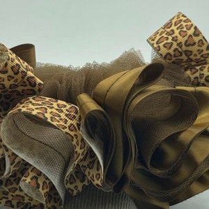 May include: A hair accessory featuring a combination of brown, tan, and leopard print ribbons. The design includes layers of ruffled ribbons and mesh, creating a textured and decorative look. The accessory is likely for special occasions.
