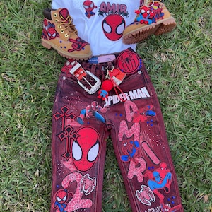 Puede incluir: Un conjunto infantil con una camiseta blanca con el nombre "AMIR" y un diseño de Spider-Man, combinado con vaqueros rojos y botas color canela, todos adornados con diseños de Spider-Man. Incluye cinturón y llavero.