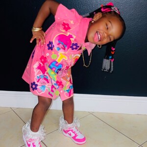 Könnte beinhalten: Ein junges Mädchen trägt ein pinkfarbenes Outfit zum Thema Trolls, bestehend aus einem kurzärmeligen Kleid und passenden Shorts. Sie trägt außerdem weiße und pinkfarbene Turnschuhe mit Rüschenmanschetten. Ihr Haar ist mit bunten Accessoires gestylt.
