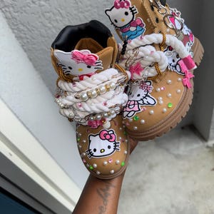 May include: Tan boots adorned with Hello Kitty patches, pink bows, and pearl accents. The laces are replaced with white rope and embellished with charms. The boots have a brown rubber sole.
