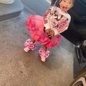 May include: A young girl wearing a white long-sleeved shirt with a pink Minnie Mouse graphic and the text "Oh Twodles" on the sleeve. She is also wearing a pink tulle skirt with white polka dots and pink and black Minnie Mouse shoes.