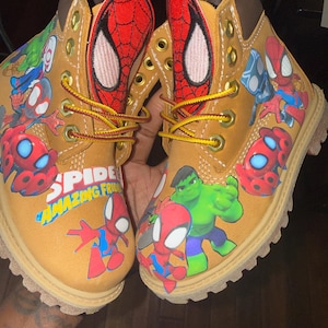 Op de afbeelding: Een paar bruine laarzen met afbeeldingen van Spider-Man en Hulk. De laarzen hebben gele veters en een bruine zool. De tekst "SPIDER AMAZING FRIENDS" is zichtbaar. Kinderschoenen.