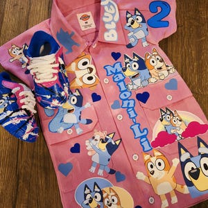 Op de afbeelding: Een roze overhemd met knopen en een blauw en wit cartoonhondontwerp. Het shirt heeft de tekst "Bluey" en "Maloni-Li" erop. Het shirt heeft ook het nummer "2" erop. Het shirt wordt gecombineerd met twee blauwe en witte sneakers.