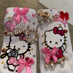 Może przedstawiać: Białe skarpetki z motywami Hello Kitty, ozdobione różowymi kokardkami, dżetami i perłowymi akcentami. Jedna skarpetka ma aparat, druga zawieszkę w kształcie gwiazdy. Zabawny i dekoracyjny dodatek.