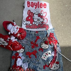 Puede incluir: Un conjunto de falda vaquera, camiseta blanca y zapatillas rojas con diseños de Hello Kitty. La camiseta blanca tiene el nombre "Baylee" y el número "4" con un gráfico de Hello Kitty. La falda tiene adornos y parches rojos.
