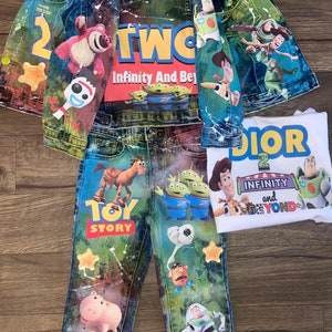 Puede incluir: Un conjunto de ropa infantil que incluye una chaqueta vaquera, unos vaqueros y dos camisetas con personajes de la película de animación "Toy Story". La chaqueta y los vaqueros están decorados con imágenes coloridas de Woody, Buzz Lightyear y otros personajes.