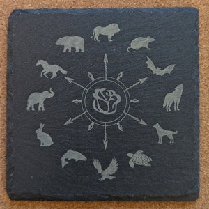 May include: A black slate coaster with a white rose design in the center. The rose is surrounded by a circle of arrows pointing outwards. Around the circle are various animal silhouettes, including a lion, bear, horse, elephant, rabbit, bat, wolf, dog, fish, turtle, and eagle.