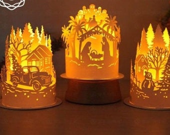Könnte beinhalten: Drei beleuchtete Papierlaternen mit filigranen Ausschnitten, die Winterszenen darstellen. Die Laternen sind weiß und zeigen einen LKW, einen Schneemann und eine Weihnachtskrippe.