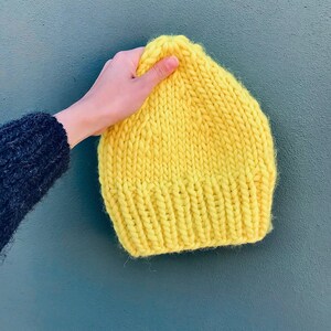 Può includere: Un berretto di lana giallo brillante. Il berretto è fatto di un filato spesso e ha un design semplice e classico.