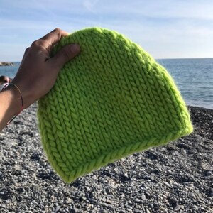Può includere: Un berretto di lana verde brillante lavorato a maglia con filato spesso. Il berretto è tenuto in una mano sullo sfondo di una spiaggia con ciottoli e l'oceano.