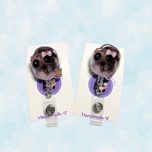 May include: Two handmade badge reels with a cartoon cat design. The cat is wearing a pink bow and has a sad expression. One cat is holding a brown and white cup. The badge reels are on white cards with purple text that says "Handmade".