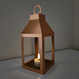 May include: A copper-colored metal lantern with a candle inside. The lantern has a square base and a rectangular frame with a pointed top. The candle is white and is lit.