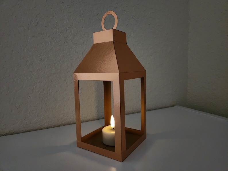 May include: A copper-colored metal lantern with a candle inside. The lantern has a square base and a rectangular frame with a pointed top. The candle is white and is lit.