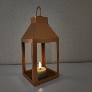 May include: A copper lantern with a candle inside. The lantern has a square base and a rectangular frame. The candle is lit and casting a warm glow.