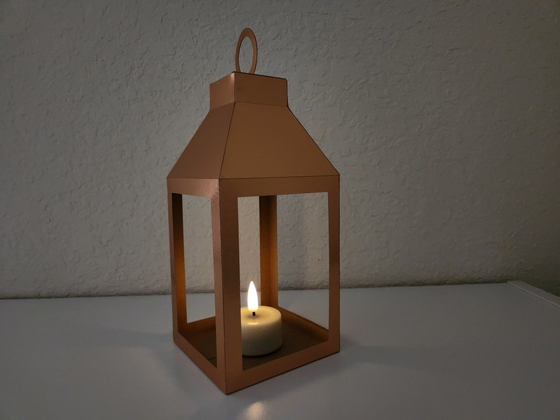 May include: A copper lantern with a candle inside. The lantern has a square base and a rectangular frame. The candle is lit and casting a warm glow.