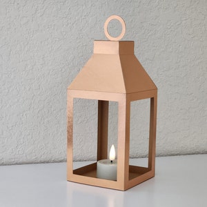 May include: A rose gold metal lantern with a candle inside. The lantern has a square base and a rectangular frame with a pointed top. The candle is a small, white tea light.