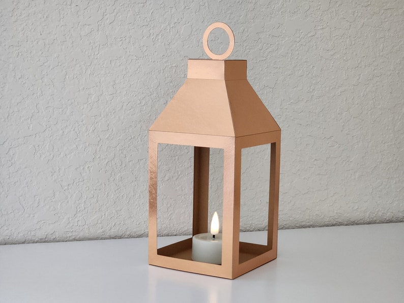 May include: A rose gold metal lantern with a candle inside. The lantern has a square base and a rectangular frame with a pointed top. The candle is a small, white tea light.