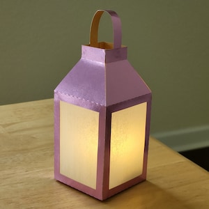 May include: A small, pink paper lantern with a white, translucent window. The lantern has a handle on the top and is lit from within.