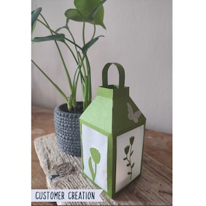 May include: A green paper lantern with a white butterfly and floral design. The lantern has a handle and is sitting on a wooden surface.
