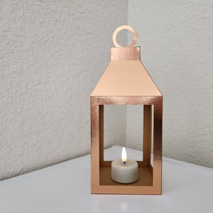 May include: A rose gold metal lantern with a candle inside. The lantern has a square base and a rectangular frame with a handle on top.