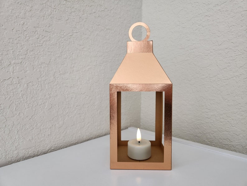May include: A rose gold metal lantern with a candle inside. The lantern has a square base and a rectangular frame with a handle on top.
