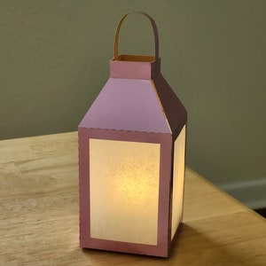 May include: A paper lantern with a light inside. The lantern is made of pink and brown paper and has a handle on top. The lantern is sitting on a wooden surface.
