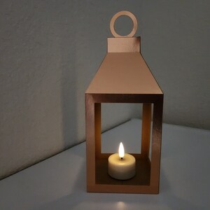 May include: A copper-colored metal lantern with a candle inside. The lantern has a square shape with a small handle on top. The candle is lit and casting a warm glow.
