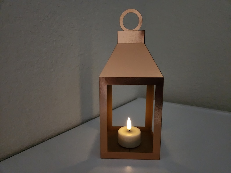 May include: A copper-colored metal lantern with a candle inside. The lantern has a square shape with a small handle on top. The candle is lit and casting a warm glow.