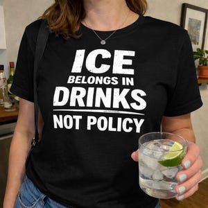 Peut inclure: T-shirt noir avec le texte blanc "ICE BELONGS IN DRINKS NOT POLICY." Un verre transparent d'eau glacée avec un quartier de citron vert est tenu dans la main. La personne porte un collier argenté.