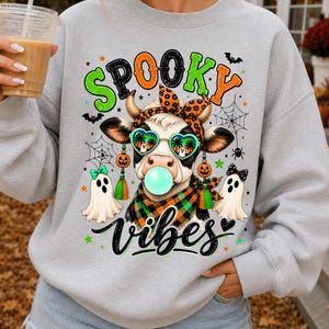 Peut inclure: Un sweat-shirt gris clair avec un motif de vache portant des lunettes de soleil en forme de cœur, un bandeau léopard et une écharpe à carreaux, faisant une bulle. Le texte "SPOOKY vibes" est entouré de fantômes, toiles d'araignée et chauves-souris.
