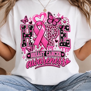 Op de afbeelding: Een wit t-shirt met een grafisch ontwerp met het woord HOPE in zwart en roze luipaardprint. Een grote roze strik, vlinders, madeliefjes en hartjes omringen de tekst. Daaronder staat BREAST CANCER AWARENESS in roze letters geschreven.