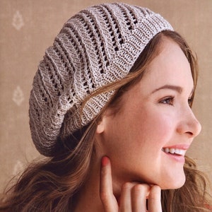 Könnte beinhalten: Eine Frau trägt eine hellbraune Strickmütze mit einem Spitzenmuster. Die Mütze ist über ihre Stirn gezogen und sie lächelt.