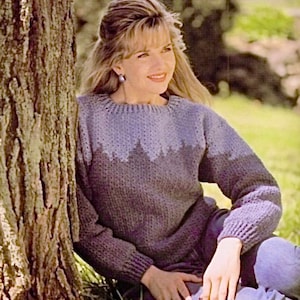Könnte beinhalten: Eine Frau trägt einen lila und blauen Strickpullover mit geometrischem Muster. Sie sitzt in einem grasbewachsenen Bereich mit einem Baumstamm hinter ihr.