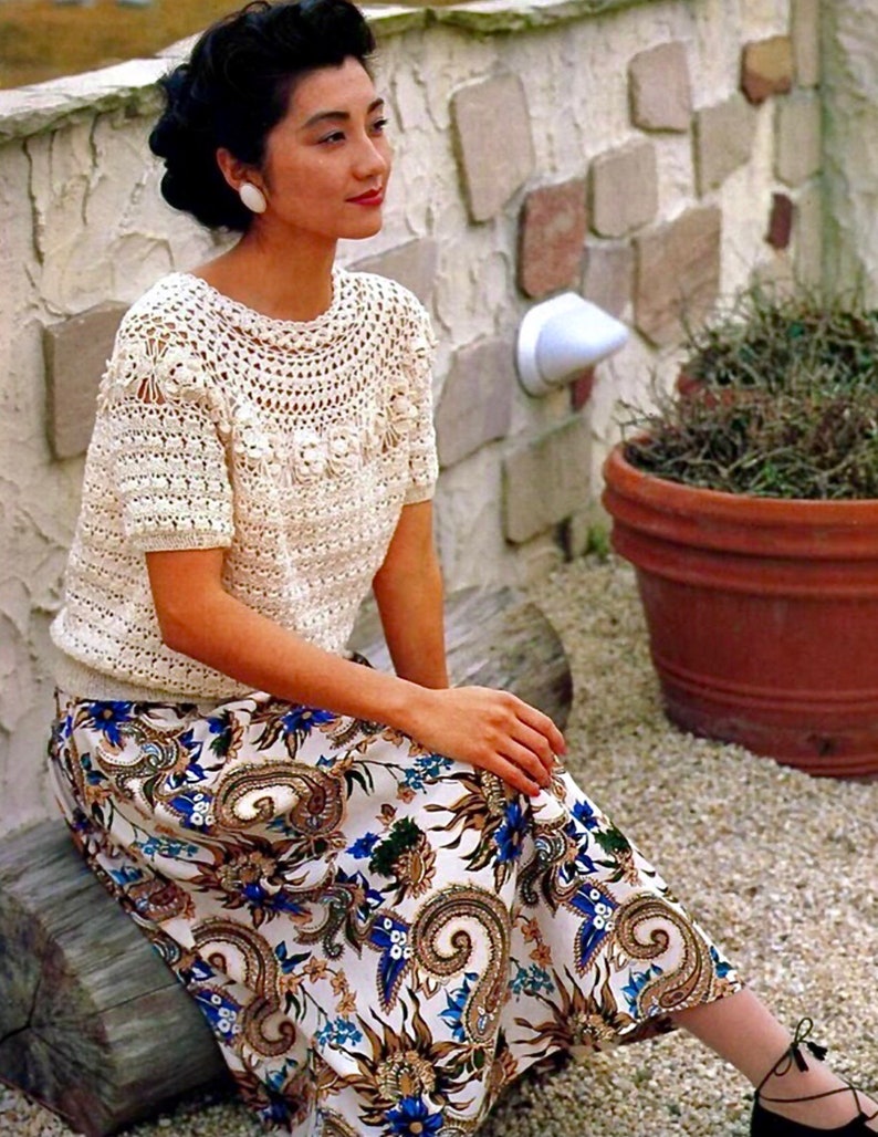 Pu&ograve; includere: Una donna indossa una maglia bianca all'uncinetto con una gonna a stampa floreale. &Egrave; seduta su un tronco di legno davanti a un muro di pietra.