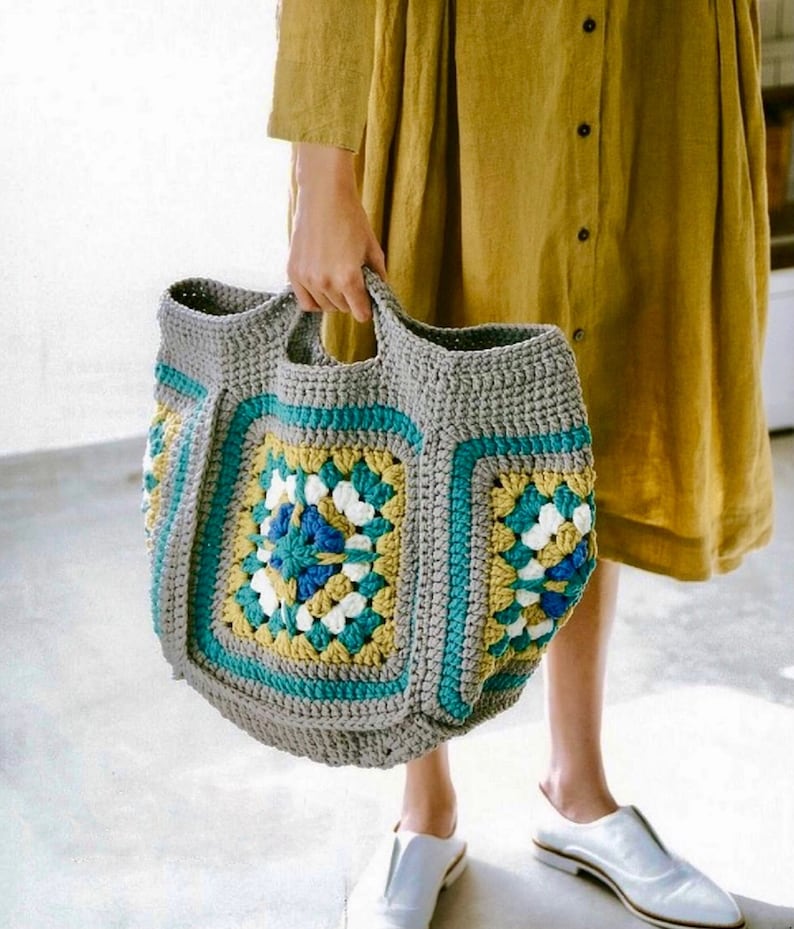 May include: A gray crocheted tote bag with colorful granny squares. The bag has two handles and is large enough to carry groceries or other items.
