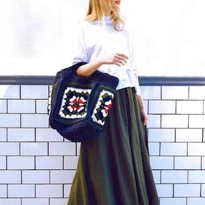 May include: A woman wearing a white long-sleeved shirt and a long green skirt is carrying a large crocheted tote bag with a colorful floral pattern. The bag is made with dark blue, green, white, red, and yellow yarn.