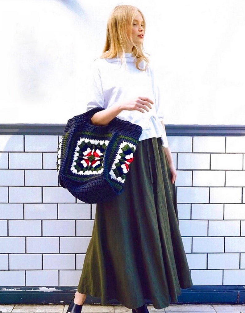 May include: A woman wearing a white long-sleeved shirt and a long green skirt is carrying a large crocheted tote bag with a colorful floral pattern. The bag is made with dark blue, green, white, red, and yellow yarn.