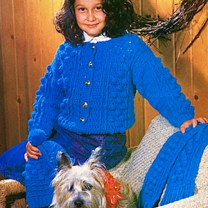 May include: A young girl wearing a blue knitted cardigan with a cable knit pattern and bobbles. She is sitting on a wicker chair with a small dog wearing a red bow.