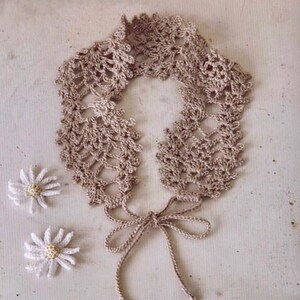 May include: A delicate crocheted collar with a lace pattern in a light brown color. The collar has a bow tie made from the same yarn.