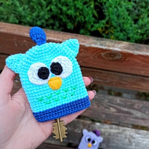 May include: A light blue crocheted owl key cover featuring a dark blue base, white eyes with black pupils, and a yellow beak. A metallic key extends from the bottom, and a small blue tuft sits on top. The owl has green stitched V-shaped details.