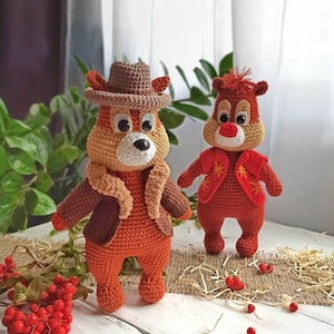 May include: Two crocheted stuffed animals, one wearing a brown cowboy hat and a brown jacket, the other wearing a red vest with a star pattern. Both are orange and brown with white faces and black eyes.