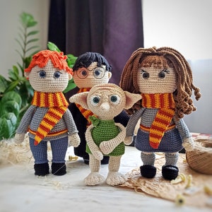 May include: Four crocheted amigurumi dolls, including Harry Potter, Ron Weasley, Hermione Granger, and Dobby the house-elf. The dolls are all wearing scarves and have detailed facial features.