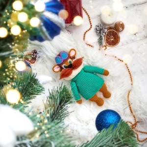 May include: A crocheted plush toy in the shape of a cartoon animal with brown fur, a green sweater, and a purple and blue hat. The toy has a red moustache and is lying on a white fluffy surface.