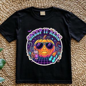 May include: A black short-sleeve t-shirt featuring a graphic of a jack-o'-lantern wearing sunglasses, holding a drink, and wearing flip-flops. The design includes the text "CREEP IT REAL" in purple letters against a retro grid background.