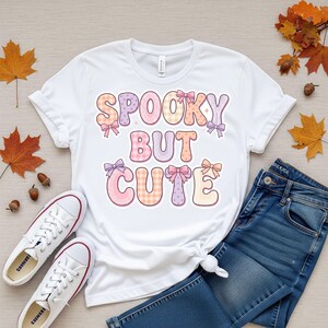 Peut inclure: Un t-shirt blanc à manches courtes avec le texte SPOOKY BUT CUTE écrit en grandes lettres arrondies aux tons pastel. Les lettres sont ornées de petits nœuds et de motifs variés, notamment du vichy, des pois et des couleurs unies rose et violet.
