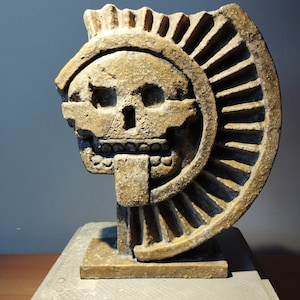 Op de afbeelding: Een bruine stenen sculptuur van een schedel met een groot, waaiervormig ontwerp erachter. Het beeld staat op een vierkante basis.