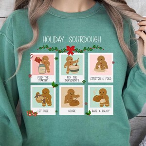 May include: A teal green sweatshirt featuring the text "HOLIDAY SOURDOUGH" and a graphic illustrating the sourdough bread-making process. The graphic shows the steps: feed the starter, mix ingredients, stretch & fold, let rise, score, and bake & enjoy.
